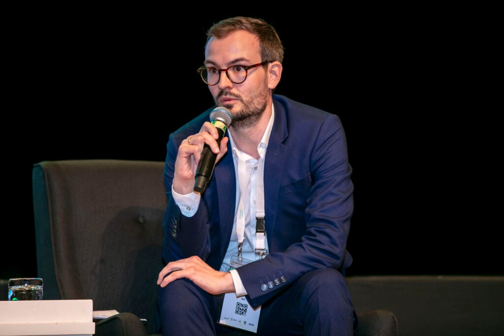 A man in a navy suit speaking into a microphone at a conference panel, with a dark background behind him.